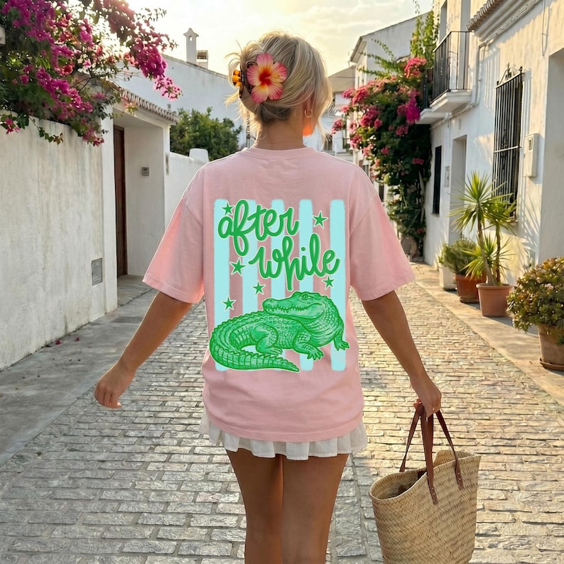 May include: A light pink t-shirt with the words "after while" in green script and a green alligator graphic. The shirt has a relaxed fit and is worn by a person walking down a cobblestone street.