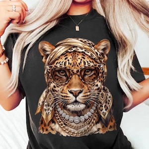Op de afbeelding: Een zwart t-shirt met een grafische print van een luipaard die een hoofddoek met luipaardprint, een cat-eye bril, bungelende gouden oorbellen en gelaagde gouden kettingen met heldere stenen draagt.