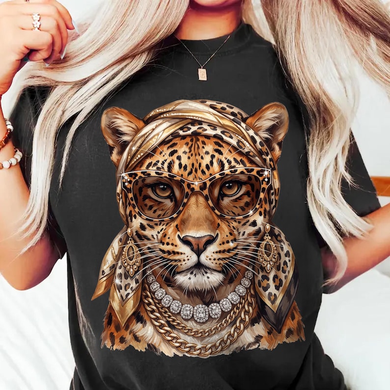 Op de afbeelding: Een zwart t-shirt met een grafische print van een luipaard die een hoofddoek met luipaardprint, een cat-eye bril, bungelende gouden oorbellen en gelaagde gouden kettingen met heldere stenen draagt.
