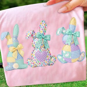 May include: Three colorful patchwork bunny decorations on a pink background. Each bunny has a different pattern, including polka dots, gingham, and floral prints. They are adorned with bows and a pom-pom tail. The bunnies are perfect for Easter.