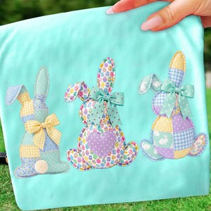 May include: A light blue fabric with three appliqued Easter bunny designs. Each bunny is made of patchwork fabric with pastel colors, polka dots, and egg patterns. The bunnies have bows and a fluffy tail.