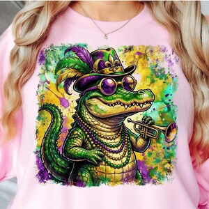 May include: Pink sweatshirt featuring a colorful illustration of a cartoon alligator wearing a hat, sunglasses, and a beaded necklace, playing a trumpet. The alligator is green, with purple, yellow, and green accents.