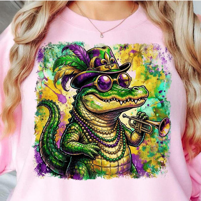 May include: Pink sweatshirt featuring a colorful illustration of a cartoon alligator wearing a hat, sunglasses, and a beaded necklace, playing a trumpet. The alligator is green, with purple, yellow, and green accents.