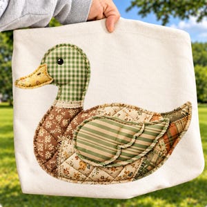 Könnte beinhalten: Eine cremefarbene Canvas-Einkaufstasche mit einer großen, gesteppten Stoffapplikation in Entenform. Die Ente besteht aus verschiedenen gemusterten Stoffstücken in Grün-, Braun- und Gelbtönen, darunter Vichy-Karo-, Blumen- und Streifenmuster.