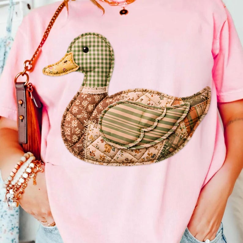 May include: Pink t-shirt featuring a patchwork duck design. The duck has a green and white checkered head, a yellow beak, and a body made of various patterned fabrics in brown, green, and beige. The duck is a whimsical, handcrafted design.