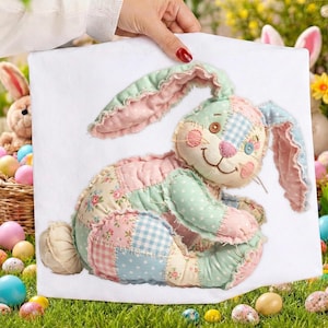 Op de afbeelding: Een patchwork konijn met roze, blauwe en groene stoffen lappen. Het konijn heeft lange oren, een knoopoog en een gestikte glimlach. Het konijn staat op een witte achtergrond, vastgehouden door een persoon.