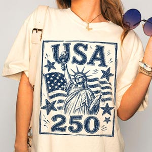 May include: Cream-colored t-shirt featuring a blue graphic of the Statue of Liberty holding a torch, with the word "USA" above and "250" below. The design includes stars and a waving American flag.
