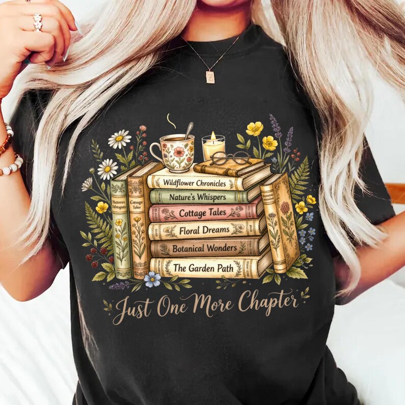 Puede incluir: Camiseta negra con un dise&ntilde;o de libros apilados adornados con ilustraciones florales y bot&aacute;nicas. Los libros tienen t&iacute;tulos como "Wildflower Chronicles" y "Botanical Wonders". Una taza de t&eacute;, gafas y una vela est&aacute;n encima. El texto "Just One More Chapter" est&aacute; debajo.