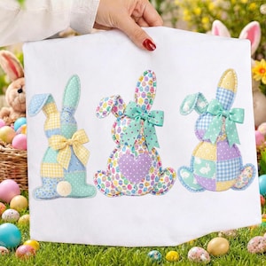 Op de afbeelding: Een witte kussensloop met drie geappliceerde paashaasjes. Elke haas heeft een patchwork-ontwerp in pastelkleuren, met strikken en decoratieve details. De kussensloop wordt door een hand omhoog gehouden.