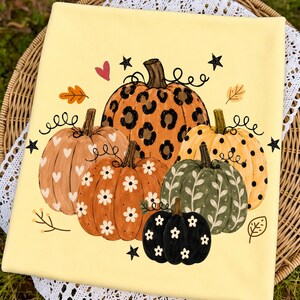 May include: A pale yellow fabric featuring a graphic print of five pumpkins. The central pumpkin has a leopard print, while others feature white hearts, white daisies, black polka dots, and white leaf patterns, accented by small black stars.