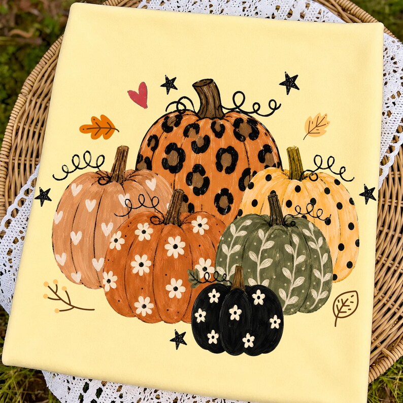 May include: A pale yellow fabric featuring a graphic print of five pumpkins. The central pumpkin has a leopard print, while others feature white hearts, white daisies, black polka dots, and white leaf patterns, accented by small black stars.