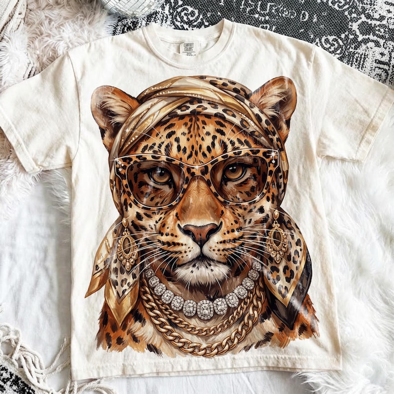 Op de afbeelding: Een cr&egrave;mekleurig T-shirt met korte mouwen en een grote grafische print van een luipaard met een hoofddoek met luipaardprint, een oversized kattenoogbril, gouden bungelende oorbellen en gelaagde gouden kettingen met een centrale kristallen hanger.