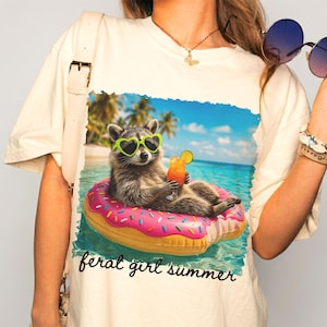 Puede incluir: Camiseta color crema con un dise&ntilde;o de un mapache con gafas de sol en forma de coraz&oacute;n flotando en una boya de donut rosa en el mar. El mapache sostiene un c&oacute;ctel. Las palabras "feral girl summer" est&aacute;n escritas en cursiva en la parte inferior.