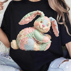 May include: Black t-shirt featuring a patchwork bunny design. The bunny is made of various pastel fabrics, including pink, blue, and green, with floral and gingham patterns. The bunny has a smiling face and button eyes. The shirt is a casual style.
