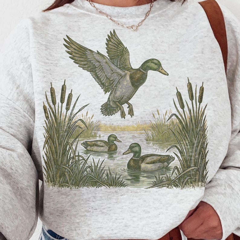 K&ouml;nnte beinhalten: Ein hellgraues Sweatshirt mit einer detaillierten Illustration von Enten in einem Teich. Eine Ente ist im Flug, w&auml;hrend zwei andere im Wasser schwimmen. Das Design umfasst Schilf und Binsen und schafft eine Natur-Thematik.