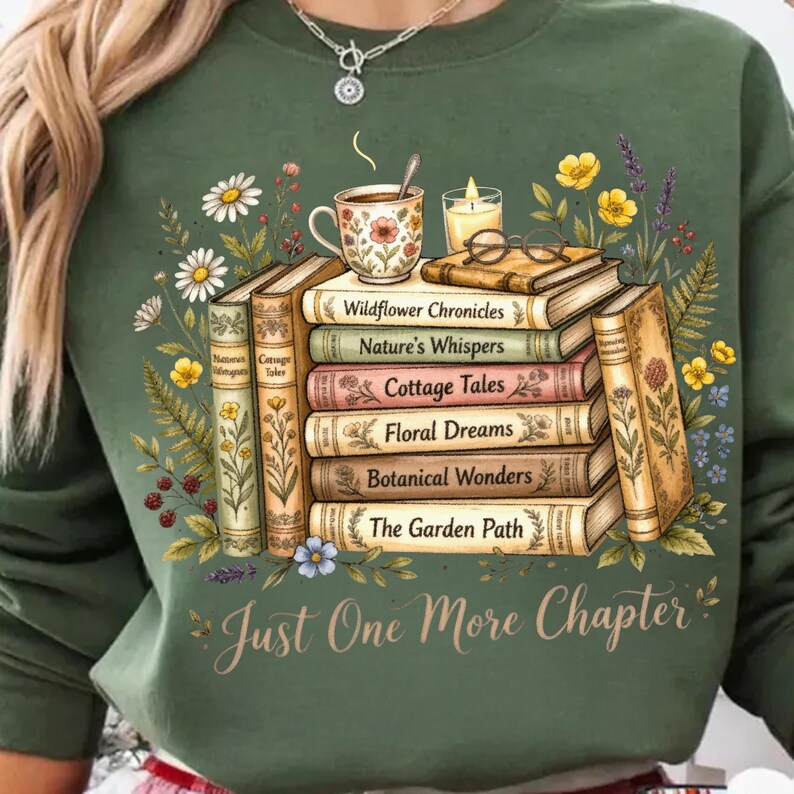 Puede incluir: Sudadera verde con un dise&ntilde;o de libros apilados, una taza de t&eacute;, una vela y gafas. Los libros tienen t&iacute;tulos como "Wildflower Chronicles" y "Botanical Wonders". El texto "Just One More Chapter" se muestra debajo, con detalles florales.