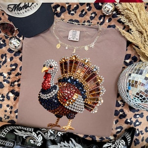 May include: A taupe t-shirt featuring a graphic of a turkey composed of multicolored rhinestones. The shirt has a Comfort Colors label at the neckline. The turkey design includes shades of red, blue, gold, and clear crystals in a fan shape.