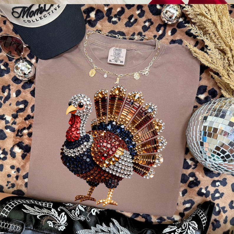 May include: A taupe t-shirt featuring a graphic of a turkey composed of multicolored rhinestones. The shirt has a Comfort Colors label at the neckline. The turkey design includes shades of red, blue, gold, and clear crystals in a fan shape.