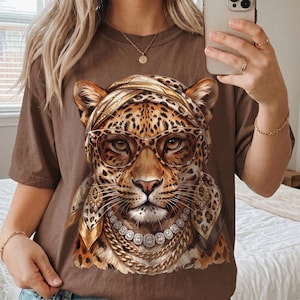 Op de afbeelding: Een bruin t-shirt met een grafische print van een luipaard met een hoofddoek met luipaardprint, een oversized bril, gouden oorbellen en gelaagde gouden en diamanten kettingen. De luipaard heeft gedetailleerde vachttexturen en een gefocuste blik.