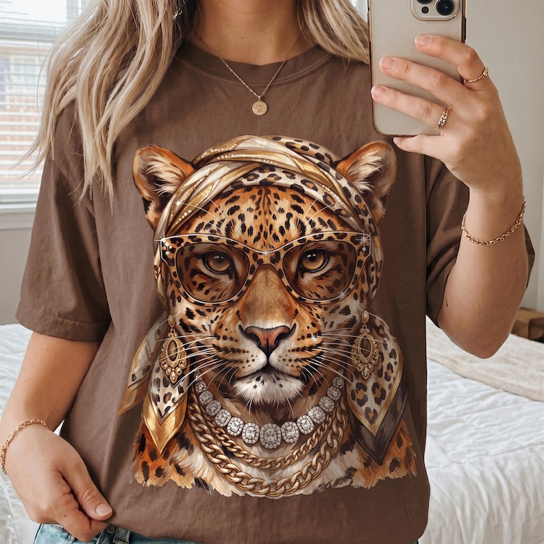 Op de afbeelding: Een bruin t-shirt met een grafische print van een luipaard met een hoofddoek met luipaardprint, een oversized bril, gouden oorbellen en gelaagde gouden en diamanten kettingen. De luipaard heeft gedetailleerde vachttexturen en een gefocuste blik.