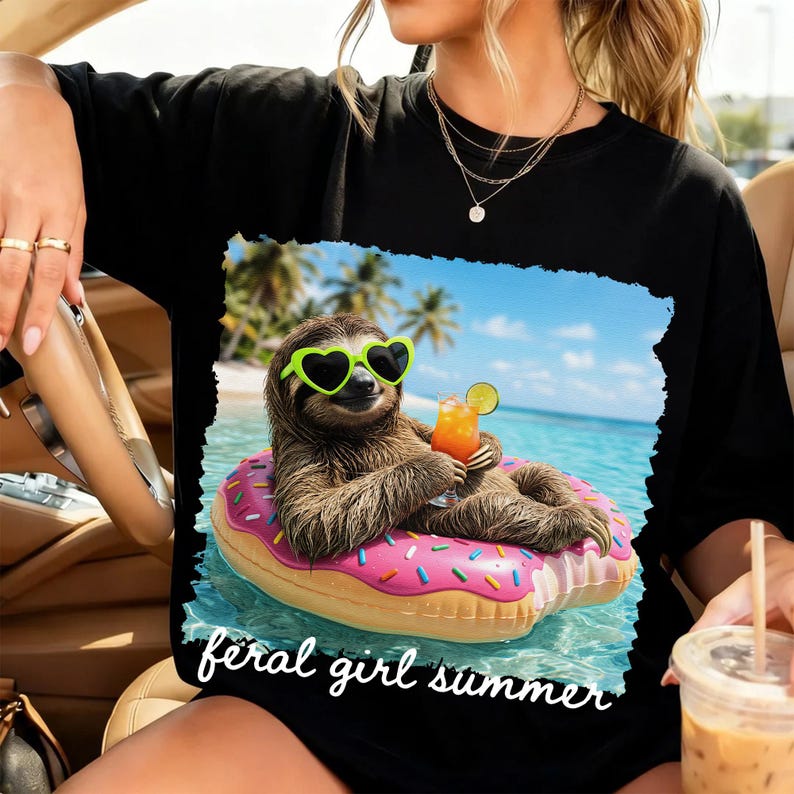 May include: Black t-shirt featuring a sloth wearing heart-shaped sunglasses and floating on a pink donut-shaped inflatable in the ocean. The sloth is holding a tropical drink. The words "feral girl summer" are written in white script.