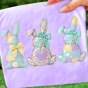 May include: Three Easter bunny appliqu&eacute;s in pastel colors on a lavender background. Each bunny is made of patchwork fabric with a bow and a heart or a pom-pom tail. The bunnies have different patterns, including polka dots, gingham, and Easter egg prints.