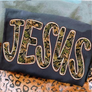 May include: Dark gray t-shirt with the word "JESUS" in large, stylized letters. The letters are filled with a camouflage pattern of brown leaves and green foliage, outlined in white. The shirt is resting on a leopard print fabric.