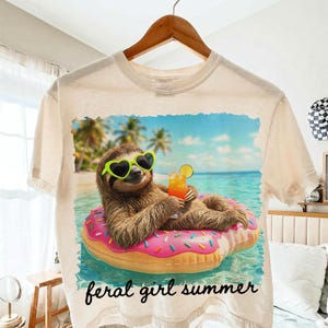 May include: Cream-colored t-shirt featuring a sloth wearing heart-shaped sunglasses and floating on a pink donut-shaped floatie in the ocean. The sloth is holding a cocktail. The words "feral girl summer" are printed in black script at the bottom.