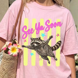 May include: Pink t-shirt featuring a raccoon illustration with outstretched arms, the text "See Ya Soon", yellow vertical stripes, and star accents. The raccoon is in shades of gray and brown.