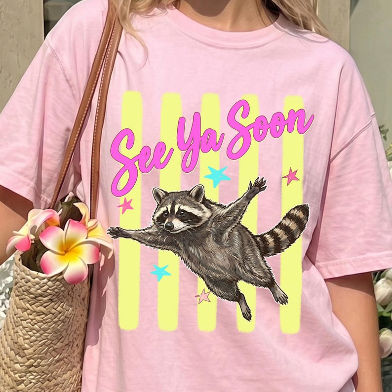 May include: Pink t-shirt featuring a raccoon illustration with outstretched arms, the text "See Ya Soon", yellow vertical stripes, and star accents. The raccoon is in shades of gray and brown.