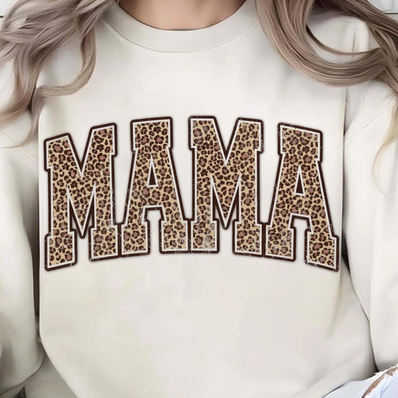 May include: Cream-colored sweatshirt with the word "MAMA" in large, block letters. The letters are filled with a brown and tan leopard print and outlined in a darker brown. The sweatshirt has a crew neck.