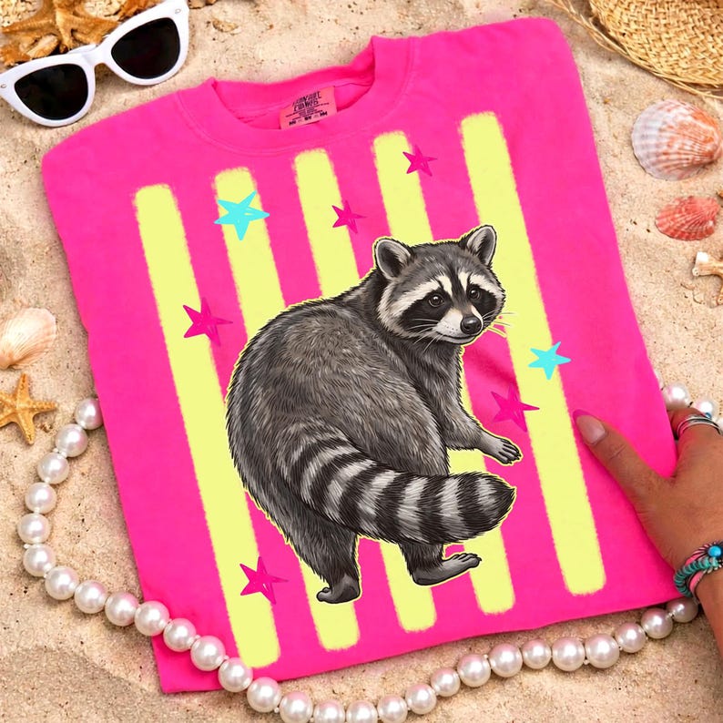 May include: A bright pink t-shirt featuring a raccoon illustration. The shirt has yellow vertical stripes and colorful star accents. The raccoon is gray and black with a striped tail. The shirt is on a sandy surface with sunglasses and a pearl necklace.