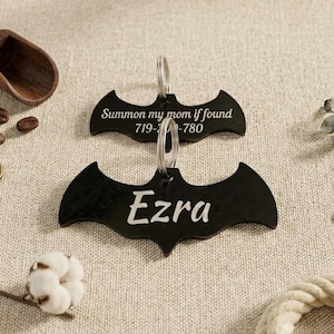 May include: Two black bat-shaped pet tags with silver key rings. The top tag reads "Summon my mom if found" followed by a phone number. The bottom tag says "Ezra" in white script. The tags are on a neutral background.