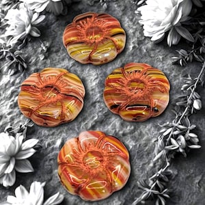 May include: Four decorative glass objects in a pumpkin shape, featuring a blend of orange, yellow, and brown colors. Each piece has a textured, swirled design and a central, textured, orange-brown detail. The objects are arranged on a gray surface.