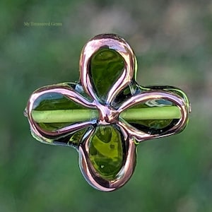 May include: A four-leaf clover-shaped pendant with a metallic rose gold frame and green glass inserts. The pendant has a glossy finish and is set against a blurred green background. The text "My Treasured Gems" is in the upper left corner.