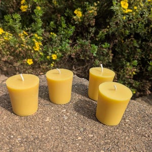 Puede incluir: Cuatro velas cilíndricas de cera de abeja amarilla con mechas blancas dispuestas sobre una superficie gris texturizada. Las velas no tienen perfume y tienen un acabado liso. El fondo presenta follaje verde y flores amarillas.