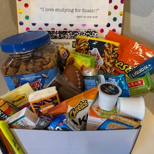 May include: A gift box filled with snacks, a football, and school supplies. The box contains a jar of Chex Mix, Cheetos, Reese's Puffs, coffee pods, and a Post-it note pad. The box has the text "I love studying for finals!!" and "- said no one ever".