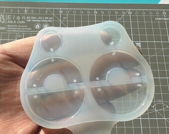 May include: A clear, flexible silicone mold with a unique shape, featuring two large circular cavities and two smaller ones. The mold is held against a gray cutting mat with centimeter markings.