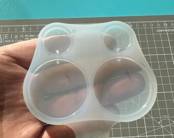 May include: A clear, flexible silicone mold with four circular cavities of varying sizes. The mold is held in a hand against a gray cutting mat with centimeter markings and a blue surface.
