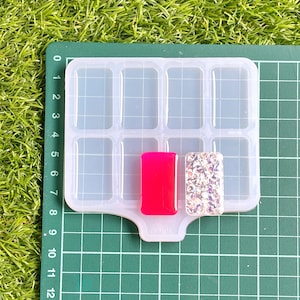 May include: A clear silicone mold with rectangular cavities, holding two rectangular resin pieces. One is bright pink, the other is covered in silver glitter. The mold sits on a green cutting mat.