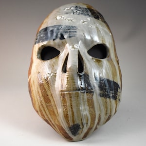 May include: A ceramic mask with a skull-like design. The mask is painted with brown, white, and black stripes and has two eye holes.