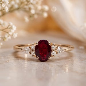 May include: A gold ring featuring a large oval-cut ruby center stone, flanked by smaller diamonds. The ring's band is a delicate gold, and the gemstones are set in a classic, elegant style. The ring is a piece of fine jewelry.