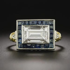 May include: A diamond ring with a rectangular central stone and a border of blue gemstones. The ring has a gold band with detailed filigree work. The central stone is surrounded by square-cut blue sapphires.