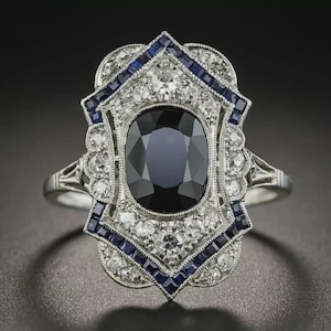 May include: An ornate silver ring featuring a large, oval, dark blue gemstone at its center. The ring is surrounded by smaller white and blue gemstones in a detailed, Art Deco-inspired design. The ring's band is also silver.