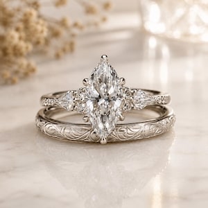 May include: A silver-toned engagement ring set featuring a large marquise-cut center stone flanked by smaller pear-shaped and round stones. The band of the second ring displays an intricate, engraved scrollwork pattern across its entire surface.