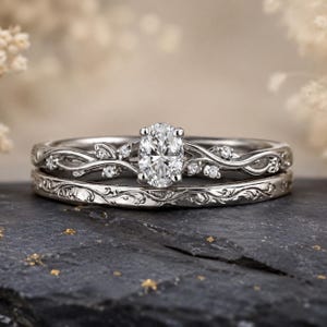 Op de afbeelding: Een zilverkleurige ringenset bestaande uit twee ringen. De bovenste ring heeft een ovaal geslepen middensteen geflankeerd door kleinere ronde stenen in een rankachtig metaalontwerp. De onderste ring is voorzien van een gegraveerd krulpatroon.