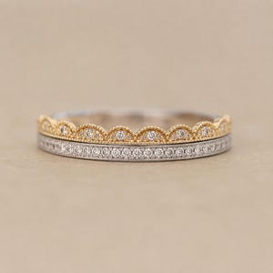 May include: Two delicate rings. The top ring is gold with a scalloped edge and small diamonds. The bottom ring is silver with a row of small diamonds. Both rings are set against a neutral background.