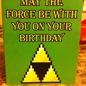 May include: A green card with the text "May the Force be with you on your birthday" in yellow letters. A yellow and black pixelated triangle is at the bottom of the card.