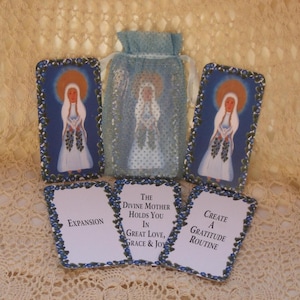 Puede incluir: Juego de cartas de oráculo con ilustraciones de una mujer con halo, en azul y blanco. El conjunto incluye una bolsa con cordón y cartas con frases como "Expansion" y "Create a Gratitude Routine".