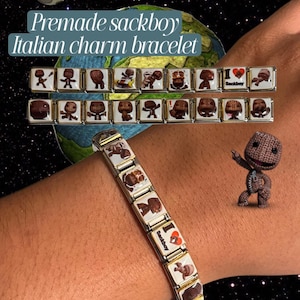 Op de afbeelding: Een zilverkleurige Italiaanse bedelarmband met meerdere vierkante schakels met bedrukte afbeeldingen van het personage Sackboy. Eén schakel toont de tekst "I heart Sackboy" met een rood hartpictogram. De armband wordt getoond om een pols.